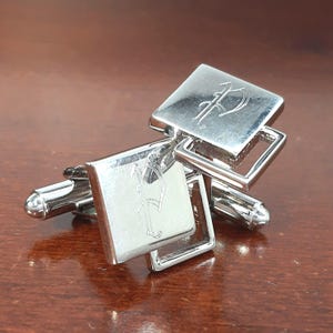 May include: Silver cufflinks with a square design. Each cufflink features a capital letter "P" engraved on the square face. The cufflinks have a polished finish and a classic, elegant style.