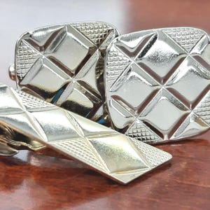 May include: A set of silver-toned cufflinks and a tie clip. The cufflinks are square with a diamond pattern, and the tie clip is rectangular with a similar design. The items are displayed on a reflective, brown surface.