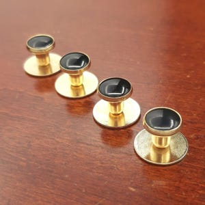 May include: Four circular studs featuring a polished gold-tone metal base and a smooth, glossy black enamel inlay on the top surface. Each stud has a short, cylindrical stem connecting the flat circular back to the rounded, black-topped head.