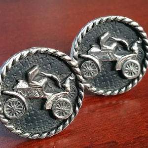 May include: A pair of silver cufflinks with a black background and a raised image of a vintage car. The car is detailed with a driver and a passenger.