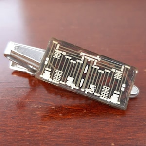 May include: A silver-toned tie clip with a rectangular, clear resin insert. The insert features a circuit board design in shades of brown, beige, and white. The tie clip is resting on a dark wooden surface.
