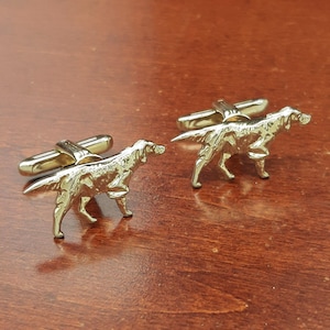 May include: A pair of metallic silver-toned cufflinks, each featuring a detailed three-dimensional figure of a standing dog with a long, flowing tail and textured coat, attached to a standard hinged toggle bar fastener for shirt sleeves.