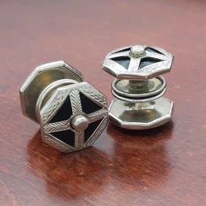 May include: A pair of silver-toned metal cufflinks featuring an octagonal shape with a central black inlay. The surface is decorated with an engraved leaf-like pattern radiating from a small central stud, and each piece has a double-disk design.