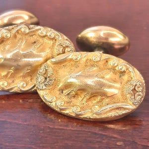 May include: Close-up of several antique gold-colored cabinet knobs. The knobs have an oval shape with ornate, detailed relief patterns. The background is a dark wood surface, providing contrast to the shiny gold finish.