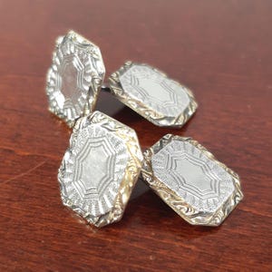 May include: Four silver cufflinks with an ornate, engraved design. Each cufflink has a rectangular shape with a detailed border and a central, patterned silver surface. The cufflinks are displayed on a dark brown wooden surface.