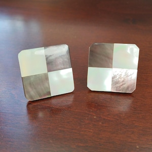 May include: Two square cufflinks with a checkerboard pattern. Each cufflink is divided into four squares, with alternating sections of iridescent white and dark gray mother-of-pearl. The cufflinks are set against a dark wood surface.