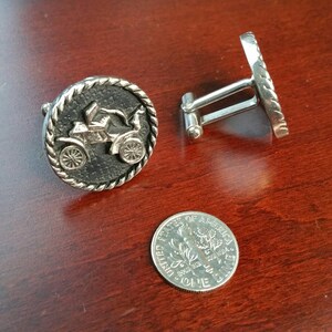 May include: A pair of silver cufflinks with a black background and a silver car design. The cufflinks are on a wooden surface.