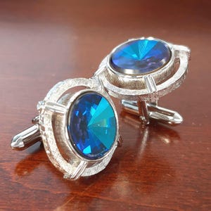 May include: Two silver cufflinks featuring large, oval, blue gemstones. The cufflinks have a textured, openwork design around the gemstones and a hinged post. The gemstones are faceted, reflecting light. The cufflinks are designed to be attached to a shirt cuff.