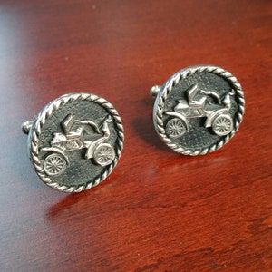 May include: A pair of silver cufflinks with a black background and a raised image of a vintage car. The cufflinks have a rope-like border.