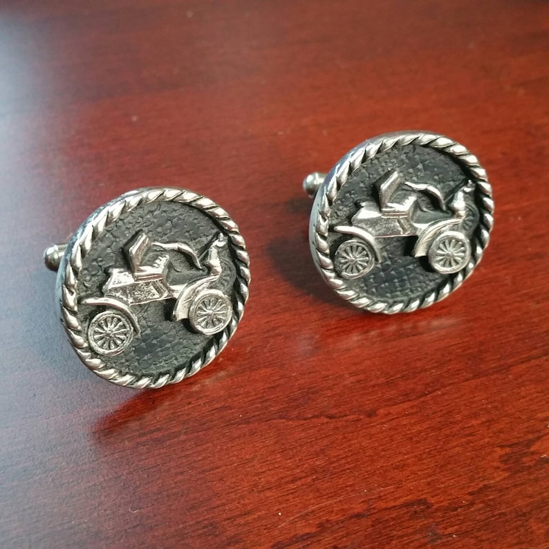 May include: A pair of silver cufflinks with a black background and a raised image of a vintage car. The cufflinks have a rope-like border.