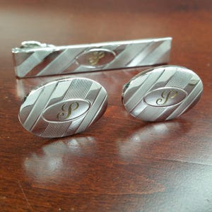 May include: A silver tie bar and cufflinks set with a striped pattern and a gold letter 'S' in the center of each piece.