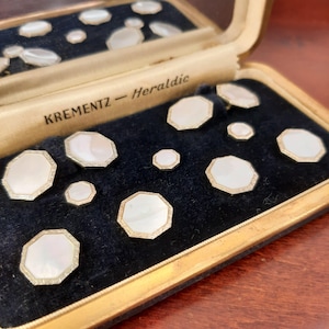May include: A collection of vintage mother-of-pearl buttons in a velvet-lined box. The buttons are octagonal with a gold-colored trim. The box is labeled "KREMENTZ - Heraldic".