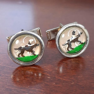 May include: A pair of silver cufflinks with a domed glass face. Each cufflink features a brown and white dog on green grass against a tan background. The cufflinks are set on a dark wood surface.