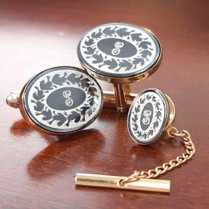 May include: A set of gold-toned metal accessories including two oval cufflinks and one smaller oval tie tack connected to a chain and bar. Each piece features a black oval center with a silver-colored letter S and a decorative silver-colored leaf border.