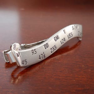 Puede incluir: Un clip de corbata de metal plateado con texto y números grabados. El clip tiene un diseño curvo con un detalle de rollo en un extremo. Los números incluyen 45, 47 ⅛, 233, 124, 183 y 256. Las letras incluyen M, RS, DD, GM, T y SOM.