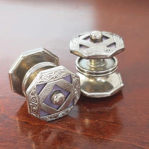 May include: Two antique silver cufflinks with a geometric design. Each cufflink features an octagonal shape with a lavender center and ornate silver detailing. The cufflinks are displayed on a dark wooden surface.