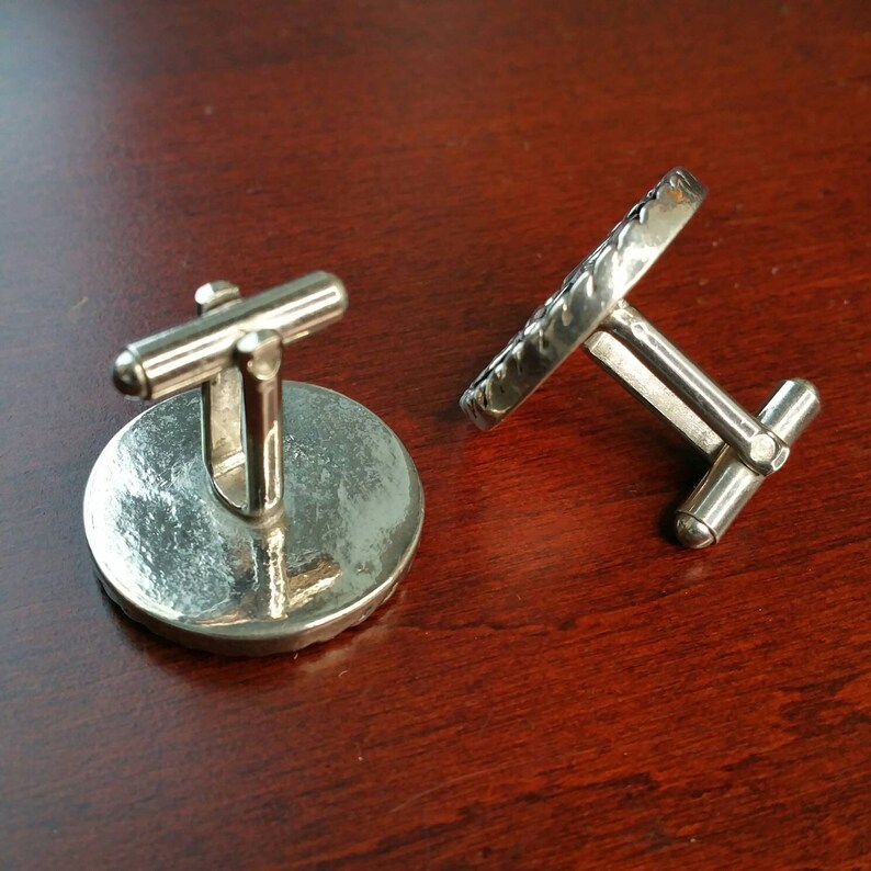 May include: A pair of silver cufflinks with a round, textured design. One cufflink has a smooth surface, while the other has a textured edge.