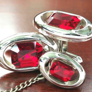 May include: A set of silver cufflinks with large, rectangular, faceted, ruby-red gemstones. The gemstones are set within an oval silver frame. A silver chain connects the two cufflinks. The cufflinks are displayed on a dark brown surface.