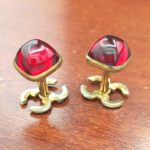 May include: A pair of cufflinks featuring square-shaped, deep red translucent glass cabochons set in gold-toned metal frames. The base of each cufflink is designed with a distinctive interlocking C-shaped metal logo motif in a gold-toned finish.