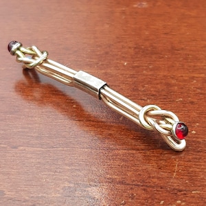 May include: A metallic tie bar featuring a central rectangular band connecting two parallel rods. Each end of the tie bar terminates in a decorative knot design, with a small, round, deep red gemstone set at the very tip of each knot.
