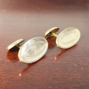 May include: A pair of vintage gold-toned cufflinks. Each cufflink features an oval face with a textured, chain-like border. The cufflinks are set against a dark brown wooden surface, showcasing their polished appearance.