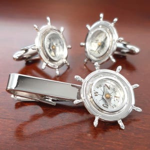 May include: A set of silver-toned cufflinks and a tie clip. Each piece features a compass design within a ship's wheel frame. The compasses have white faces with black lettering and a gold-colored needle.