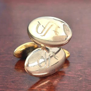 May include: Gold-toned oval cufflinks with engraved initials. The cufflinks are highly polished, reflecting light, and are arranged on a dark wooden surface. The initials are in a script font, adding a touch of elegance to the accessory.