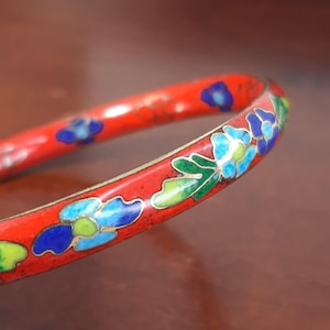 Puede incluir: Brazalete de esmalte rojo con diseño floral. El brazalete presenta flores azules, verdes y blancas con detalles dorados. Una pieza de joyería clásica.