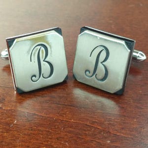 May include: A pair of square silver-toned cufflinks featuring a dark, engraved script letter B centered on each face. The cufflinks have a polished metallic finish with a dark border and a standard metal toggle mechanism on the back for fastening.