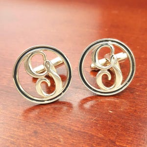 May include: A pair of circular silver-toned cufflinks featuring an openwork design with a stylized letter G in the center. Each cufflink has a polished metallic finish with a thin black circular border framing the central letter detail.