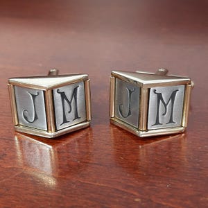 Könnte beinhalten: Ein Paar silber-goldene Manschettenknöpfe. Jeder Manschettenknopf ist würfelförmig und trägt die Buchstaben "J" und "M" in Schwarz. Die Kanten sind goldfarben, die Flächen silbern. Die Manschettenknöpfe liegen auf einer braunen Oberfläche.