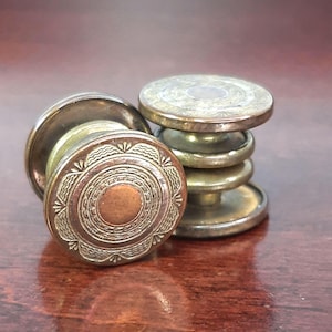 May include: Two antique brass cabinet knobs with decorative circular designs. One knob is lying on its side, revealing a copper-colored center. The other knob is stacked with several similar pieces, showcasing the hardware's construction.