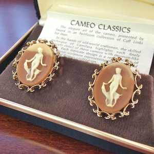 May include: Two gold-tone cameo cufflinks with a brown and white cameo design of a woman holding a bird. The cufflinks are in a velvet box with the text "CAMEO CLASSICS" and a description of the craftsmanship.