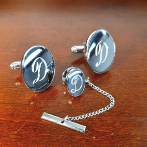 May include: A set of silver-toned cufflinks, tie tack, and shirt studs. Each piece has a black oval face with a cursive letter "D" in white. The tie tack is connected to a chain and bar.