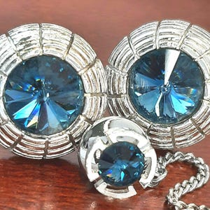 Könnte beinhalten: Ein Set aus Vintage-Schmuck mit einem Paar runder Ohrringe und einem passenden Anhänger. Die Ohrringe und der Anhänger haben eine silberfarbene Metalleinfassung mit geripptem Design, die große, facettierte blaue Kristalle umgibt. Eine silberne Kette ist am Anhänger befestigt.