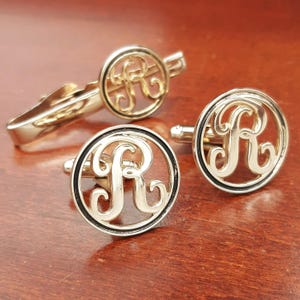 May include: A gold-toned tie bar and a pair of matching cufflinks, each featuring a circular frame with an ornate, stylized letter R monogram in the center. The metal has a polished finish, and the circular designs are consistent across all pieces.