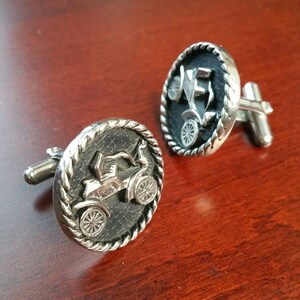 May include: A pair of silver cufflinks with a vintage car design. The cufflinks have a black background with a silver car design. The car is in a raised design.