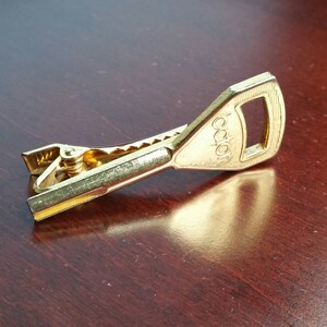Puede incluir: Un broche para corbata de color dorado con forma de llave y la palabra "Japan" grabada en la parte superior.