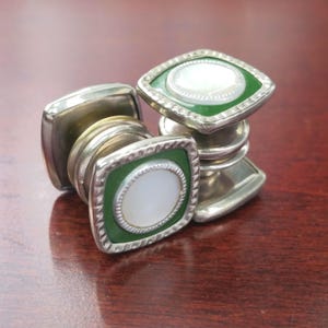 May include: A pair of vintage cufflinks with a silver finish. Each cufflink features a square green enamel border surrounding a white, oval-shaped center. The cufflinks are designed with a classic, elegant style.