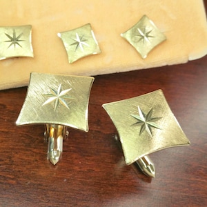 May include: Gold-toned cufflinks with a starburst design. The cufflinks have a square shape with a textured surface and a starburst pattern in the center. The image shows several cufflinks, some laid out on a beige surface and others on a dark wooden surface.