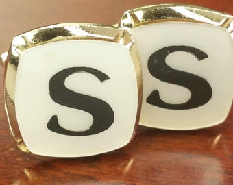 May include: A pair of gold-toned cufflinks featuring a square shape with rounded corners. Each cufflink has a white face with a bold, black capital letter S centered on the surface. The metallic edges are polished and reflective.