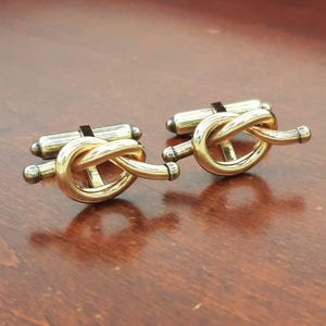 Pode incluir: Um par de abotoaduras de metal em tom dourado com um design de nó polido. Cada abotoadura possui um laço liso e arredondado em forma de nó, fixado a um mecanismo de barra basculante de metal em tom prateado para prender no punho.
