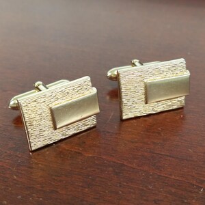 May include: A pair of gold-toned cufflinks with a textured rectangular design. Each cufflink has a smaller rectangular inset.
