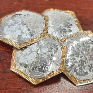 May include: Four silver-toned cufflinks with gold-colored edges. Each cufflink has a floral design in the center. The cufflinks are arranged on a brown surface.