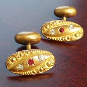 Pode incluir: Um par de botões de punho dourados com um design oval. Cada botão de punho apresenta uma pedra vermelha central ladeada por duas pedras transparentes, inseridas num padrão ornamentado e gravado. Os botões de punho têm um estilo clássico e elegante.