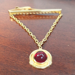 May include: A gold-coloured tie clip with a chain and a pendant. The pendant features a round, red gemstone set in a gold frame. The tie clip is designed to secure a tie to a shirt.