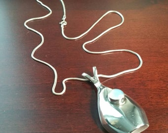 Puede incluir: Un collar de cadena de plata con un colgante con forma de pez. El pez tiene una piedra redonda blanca en su vientre.