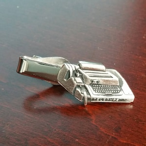 May include: A silver tie clip shaped like a vintage Teletype machine. The clip has a detailed design with a keyboard and the word "Teletype" engraved on the side.