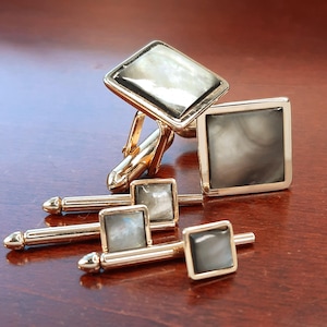 May include: A set of gold-toned cufflinks and shirt studs. The cufflinks have rectangular and square mother-of-pearl inlays. The shirt studs have square mother-of-pearl inlays. The set is displayed on a dark wood surface.