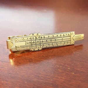 Könnte beinhalten: Ein goldfarbener Rechenschieber im Vintage-Stil, ein mathematisches Instrument zur Berechnung. Der Rechenschieber hat eingravierte Zahlen und Markierungen, eine rechteckige Form und ein verschiebbares Mittelteil. Das Instrument wird auf einer Holzoberfläche gezeigt.